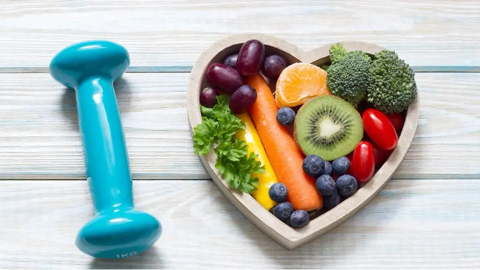 A heart shaped bowl with fruits and vegetables in it next to a blue dumbbell